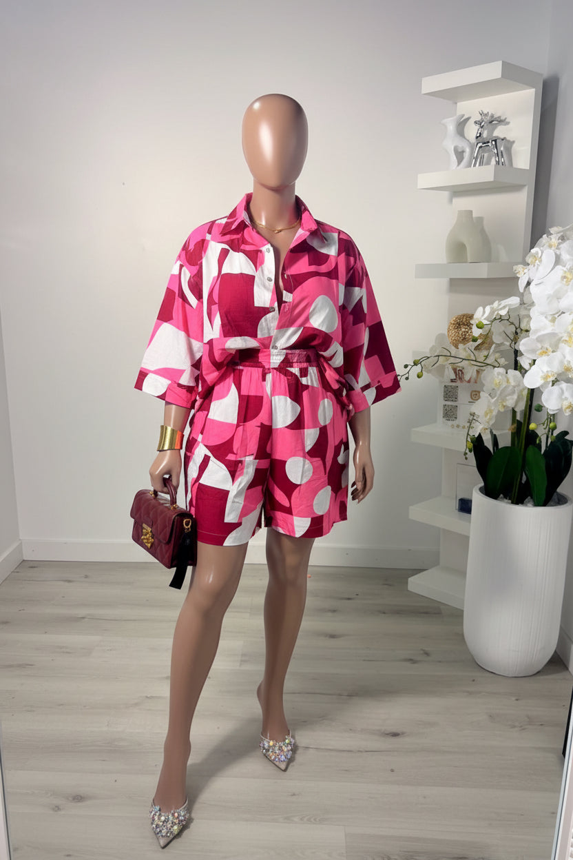 Woman taking a mirror selfie wearing a pink and white patterned dress in a room with a shelf and flowers.